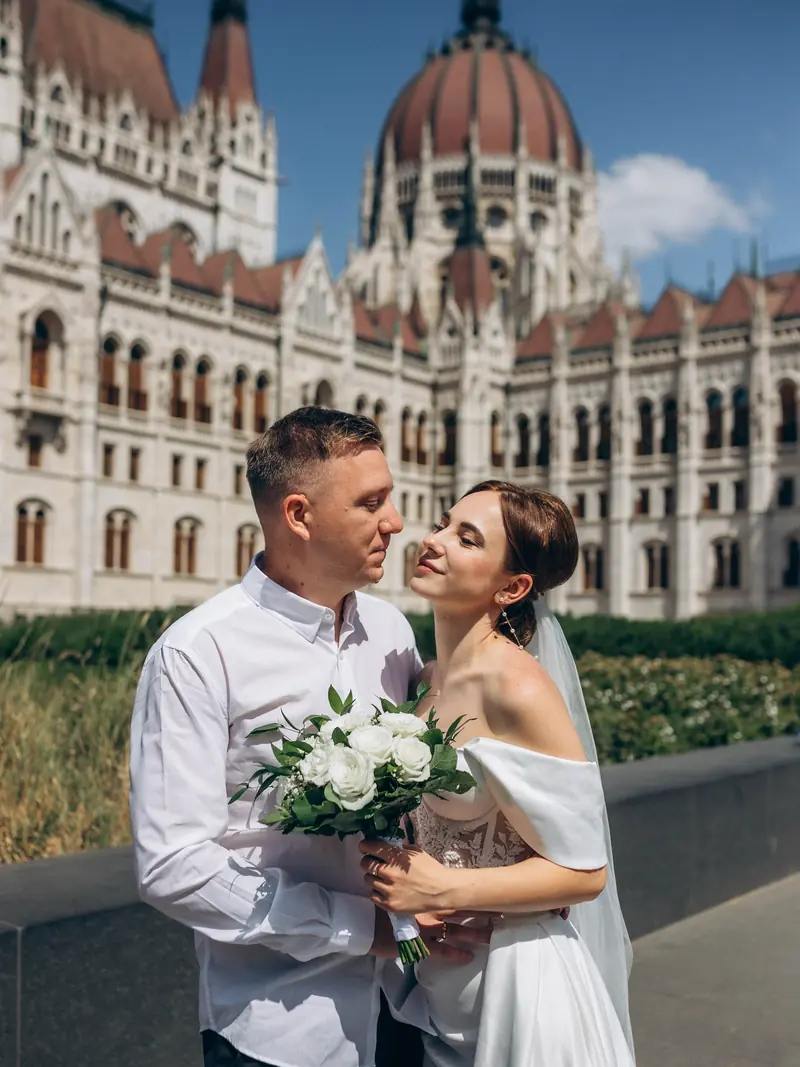 Newlyweds embracing in front of the red dome and spires of the Hungarian Parliament under a blue sky.