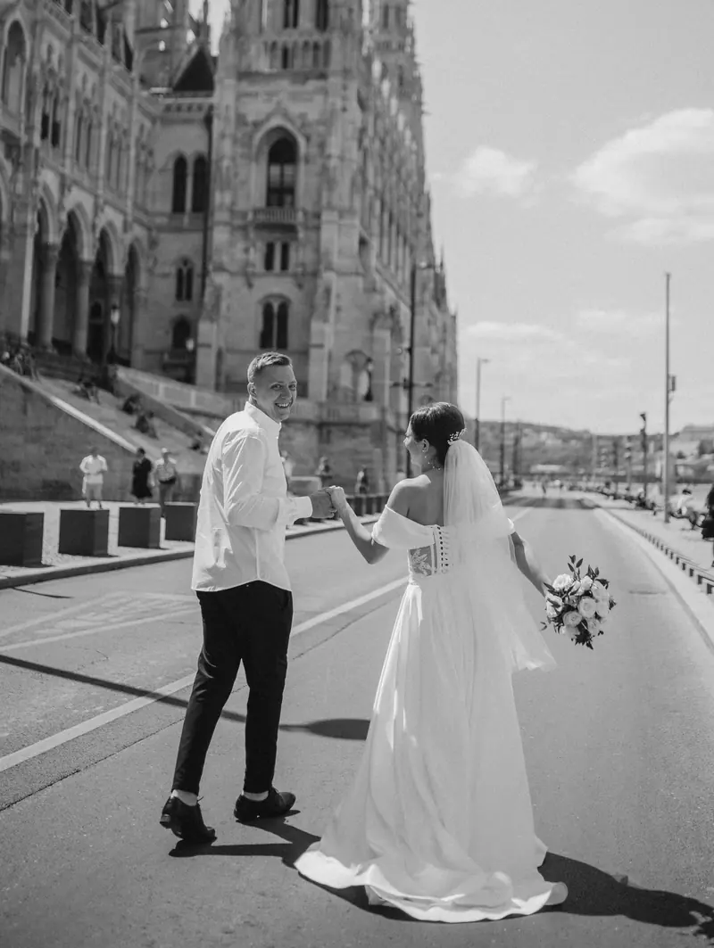 Black-and-white candid of newlyweds walking hand in hand beside the Hungarian Parliament.