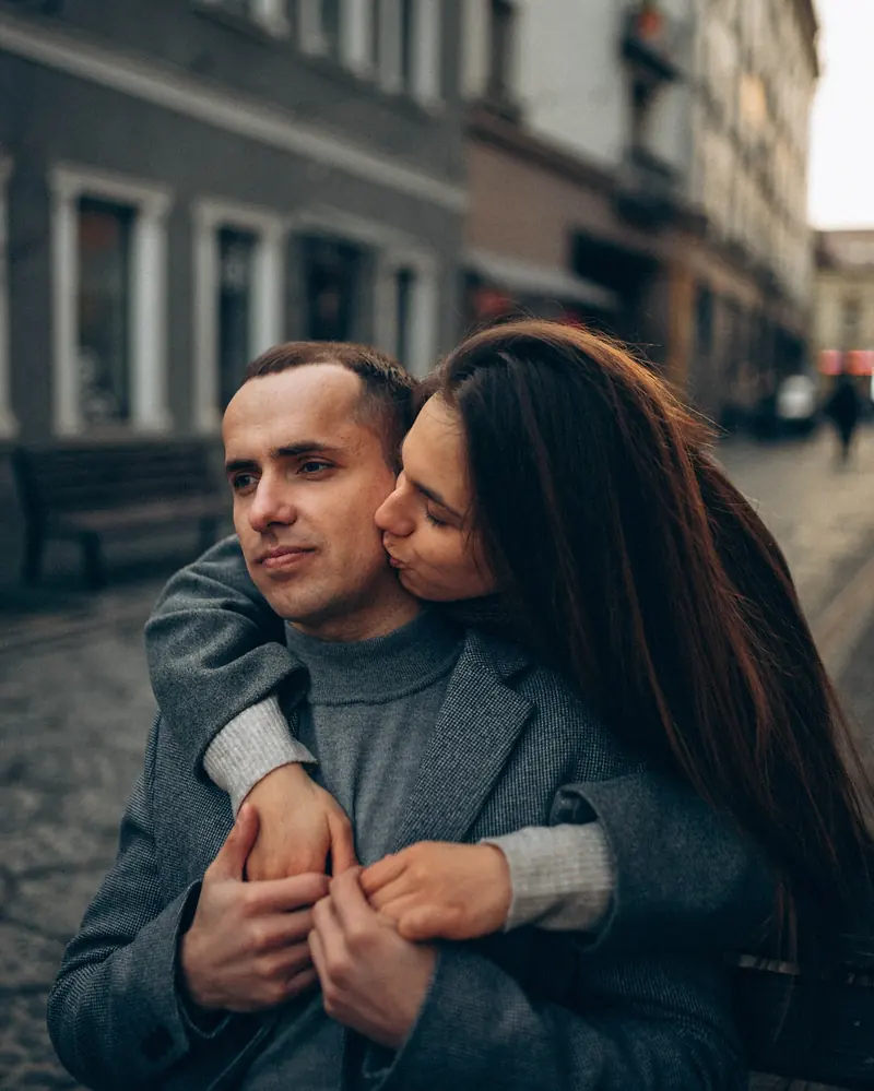 She hugs him from behind and kisses his cheek on an old-town street.