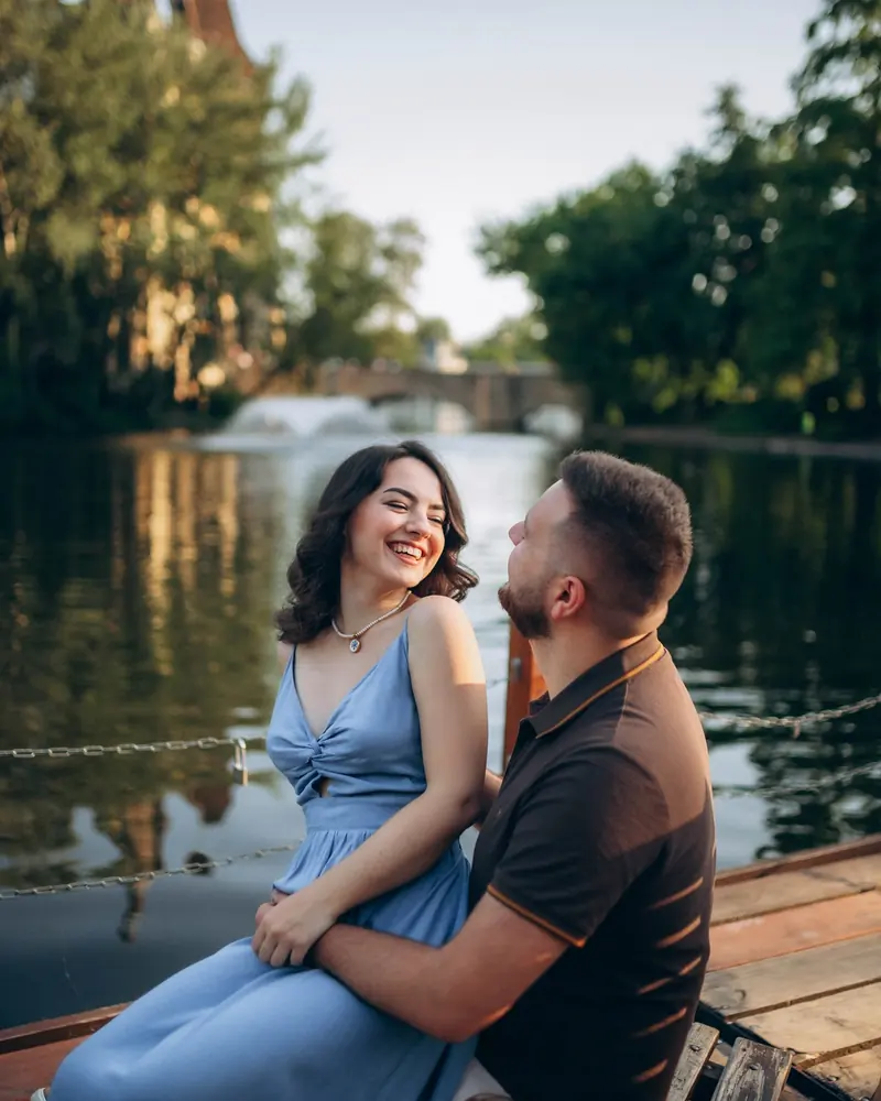Couple laughing together on a wooden lakeside bench in Budapest's City Park, warm summer evening light.