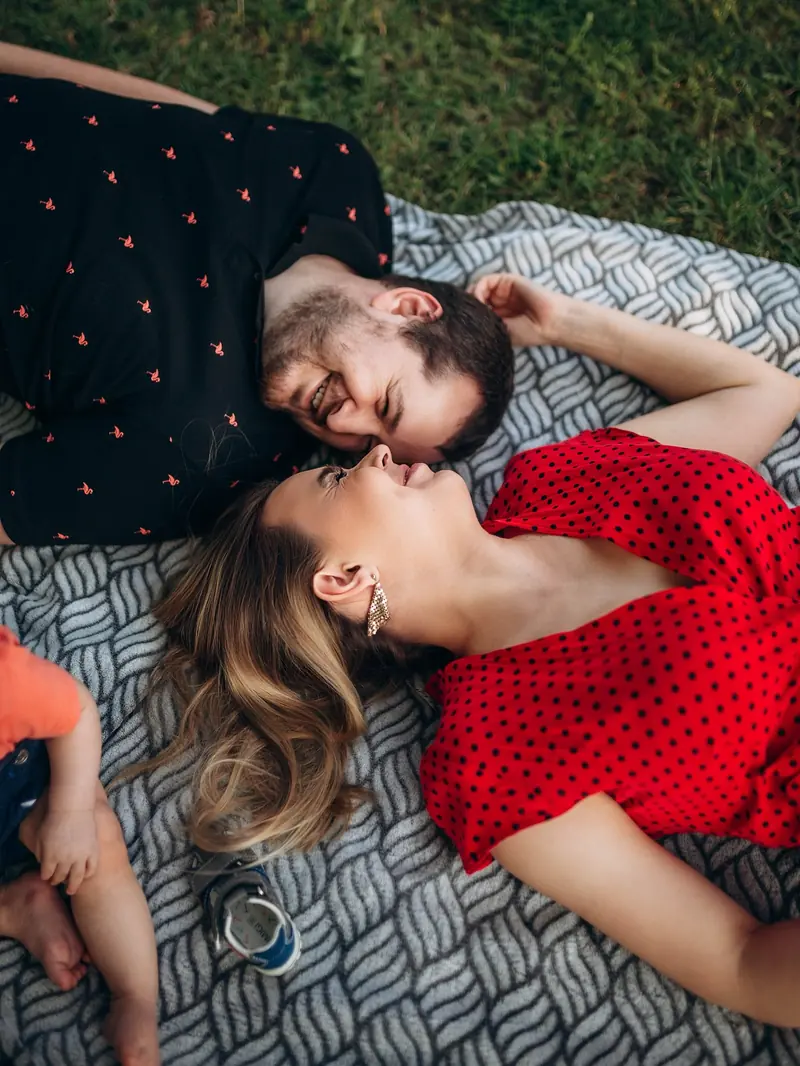 Parents lying on a picnic blanket in City Park, nose to nose and laughing, their baby's feet just in the frame.
