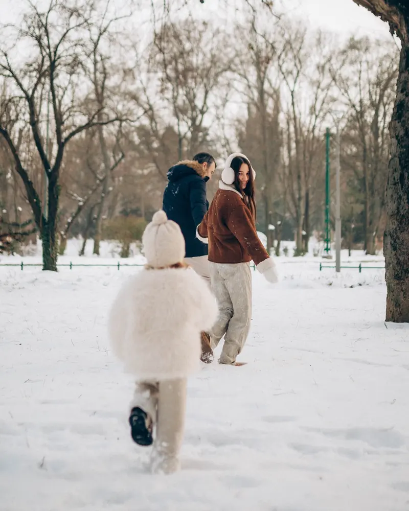 A toddler in a white fur coat runs through the snow after mum and dad in a Budapest park, seen from behind.