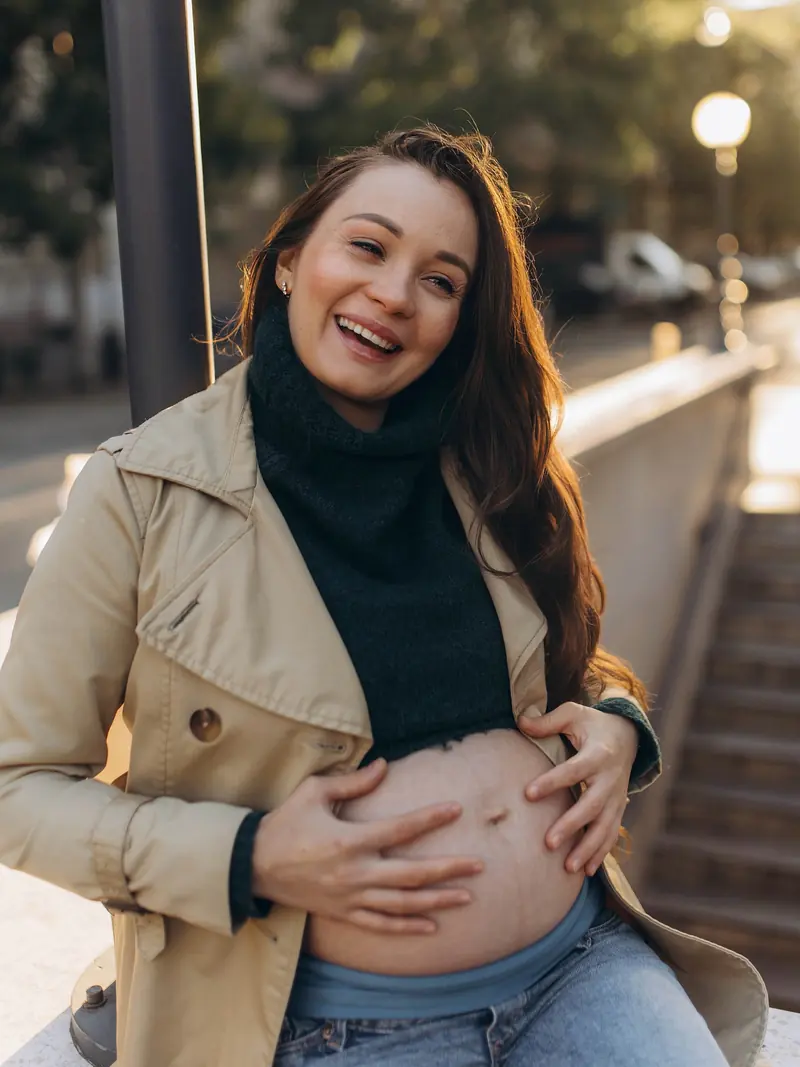 Mother-to-be in a beige trench coat laughs with both hands on her bare bump on a Budapest street at golden hour.