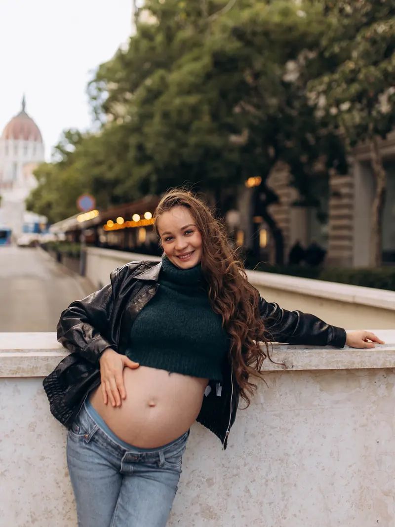 Smiling expectant mother in a leather jacket and green knit, one hand on her bump, leaning on a stone wall on a Budapest street with the Parliament dome behind her.