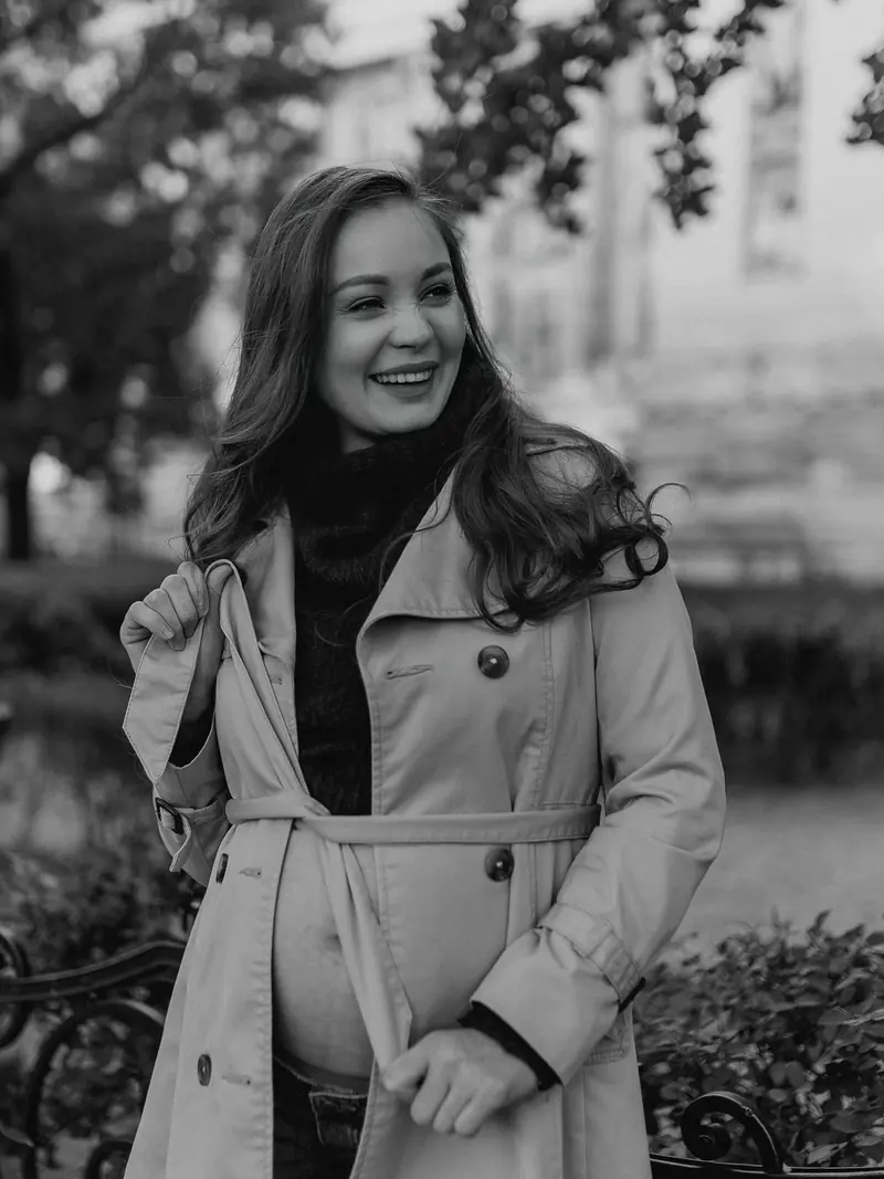 Black-and-white portrait of the expectant mother in a belted trench coat, hands resting on her bump.