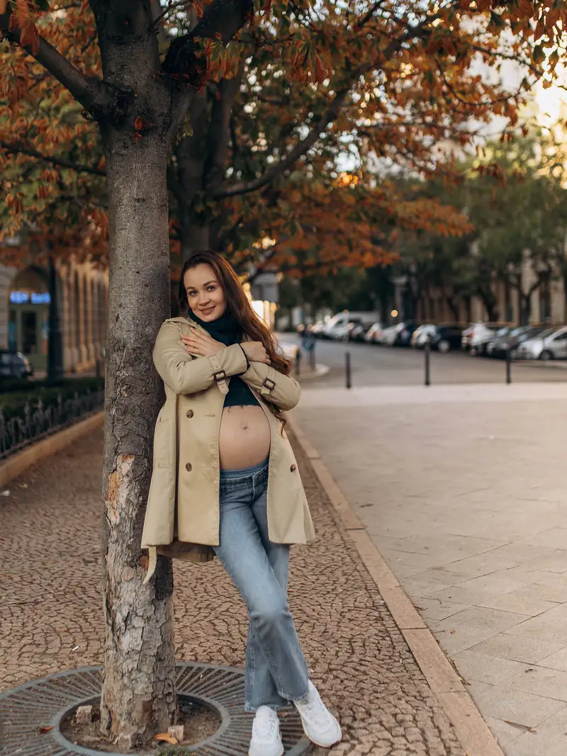 Full-length autumn portrait under a tree with orange leaves, trench coat open over the bump.