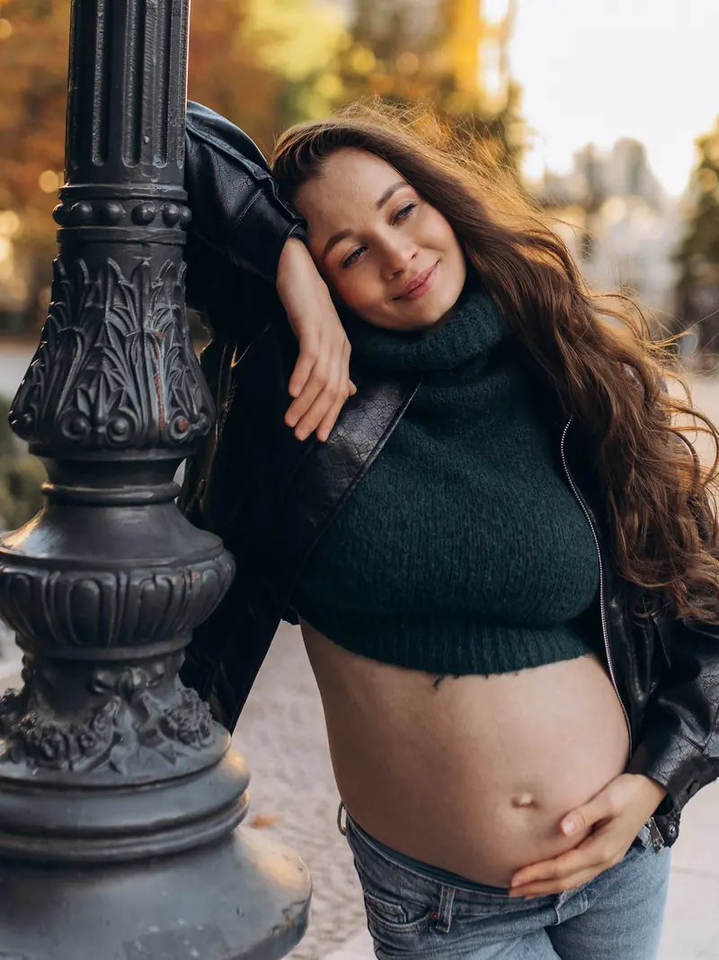 Relaxed pose against an ornate street lamp, eyes closed and smiling, hand on her bump.