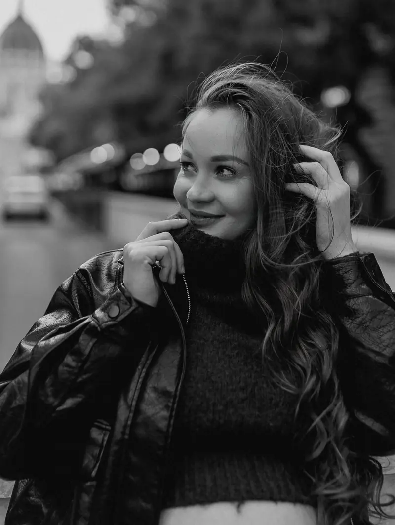 Black-and-white candid of the mother-to-be in a leather jacket, hand in her hair, on a quiet street.