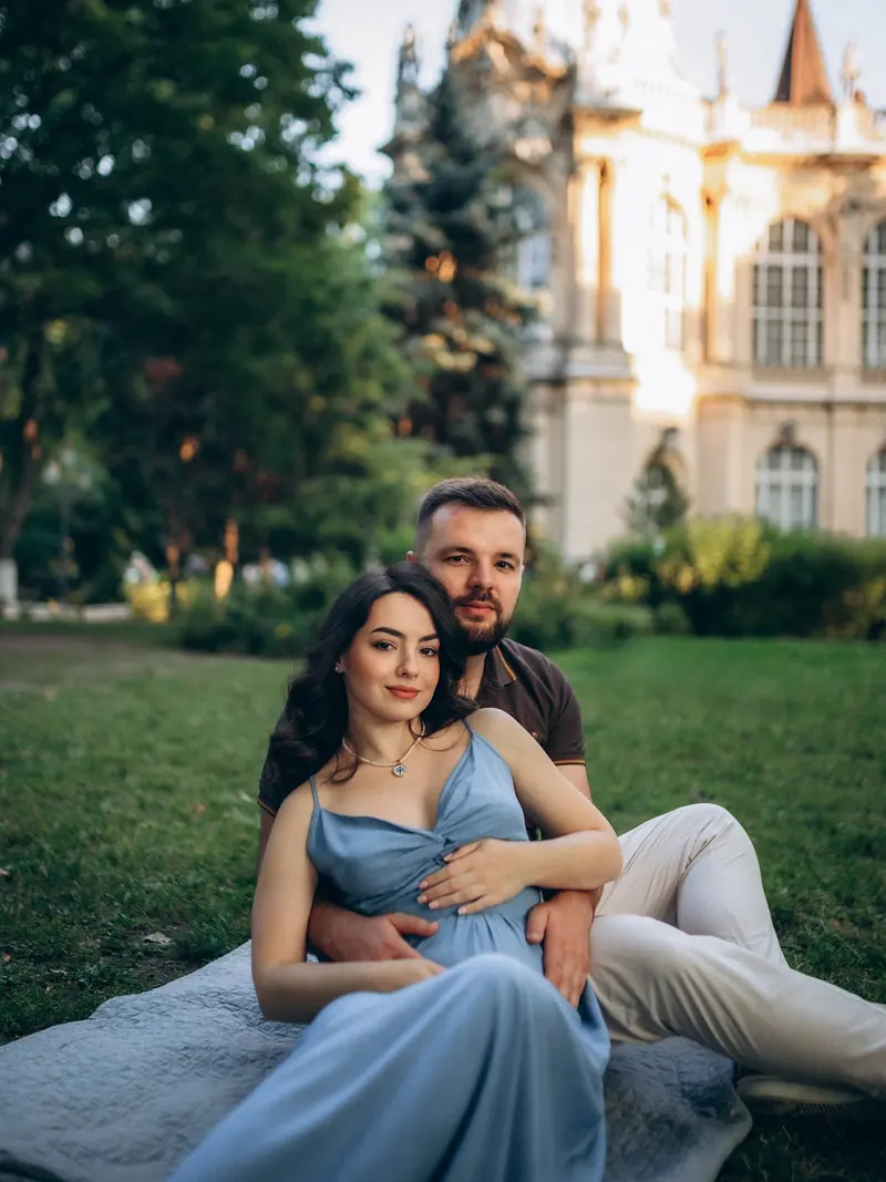 Expecting couple sitting together on a blanket in City Park, his arms around her and her bump, Vajdahunyad Castle behind them.