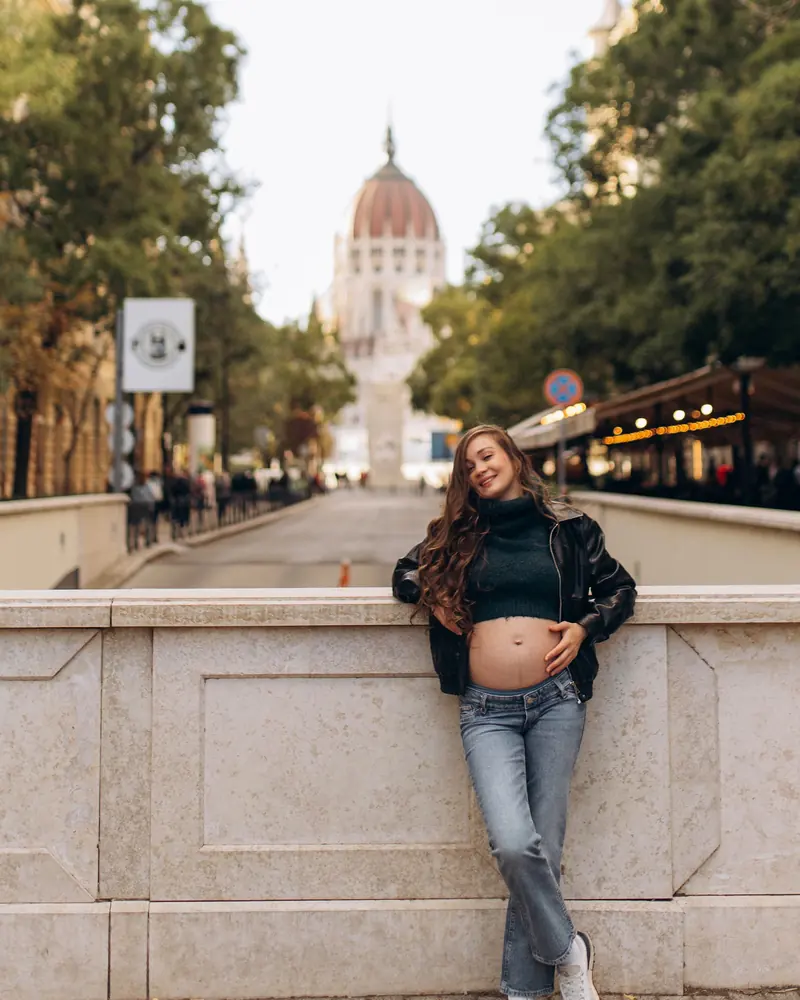Expectant mother in a leather jacket and green knit leans on a stone balustrade, one hand on her bump, with the Parliament dome down the street behind her.