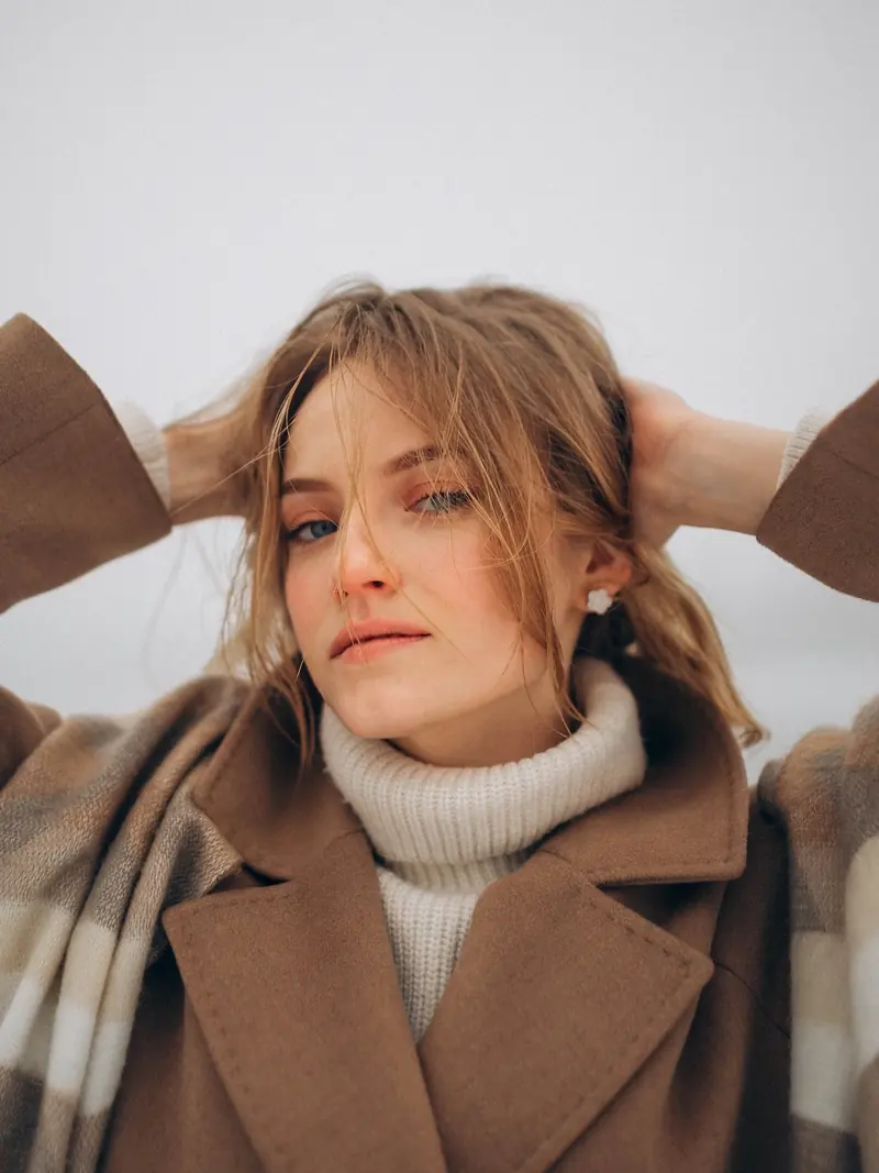 Winter portrait by the Danube in a camel coat and white turtleneck, hands in her hair.