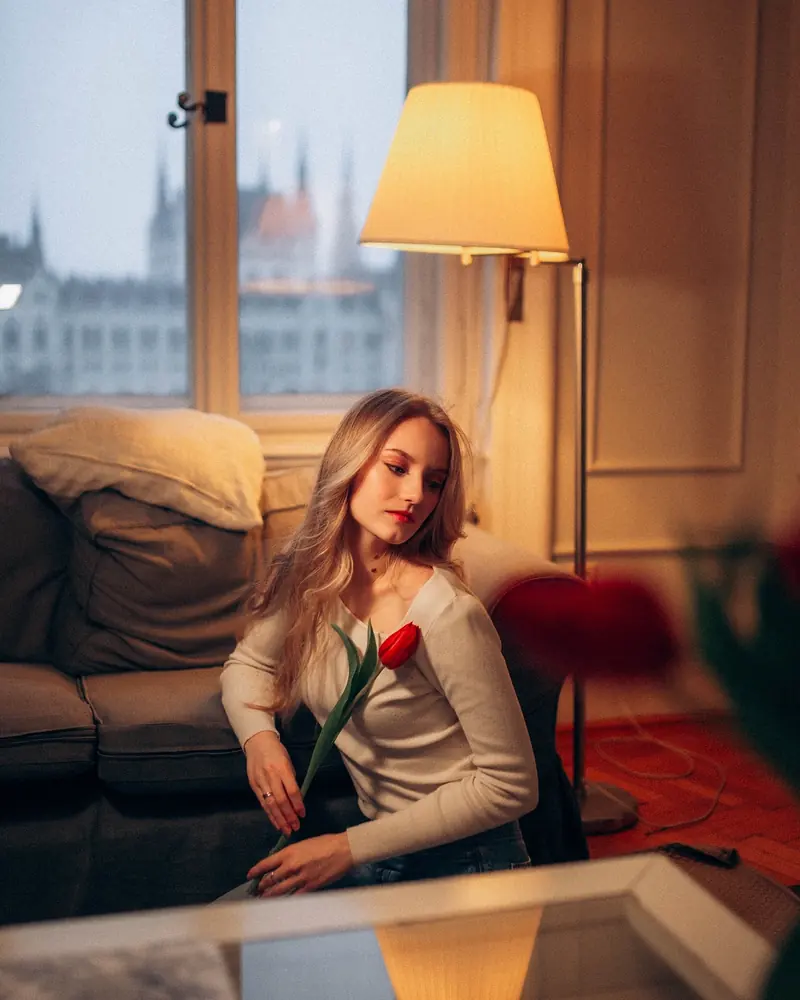 Woman with a red tulip seated by a warm lamp in a Budapest apartment, the Parliament dome framed in the window behind her.