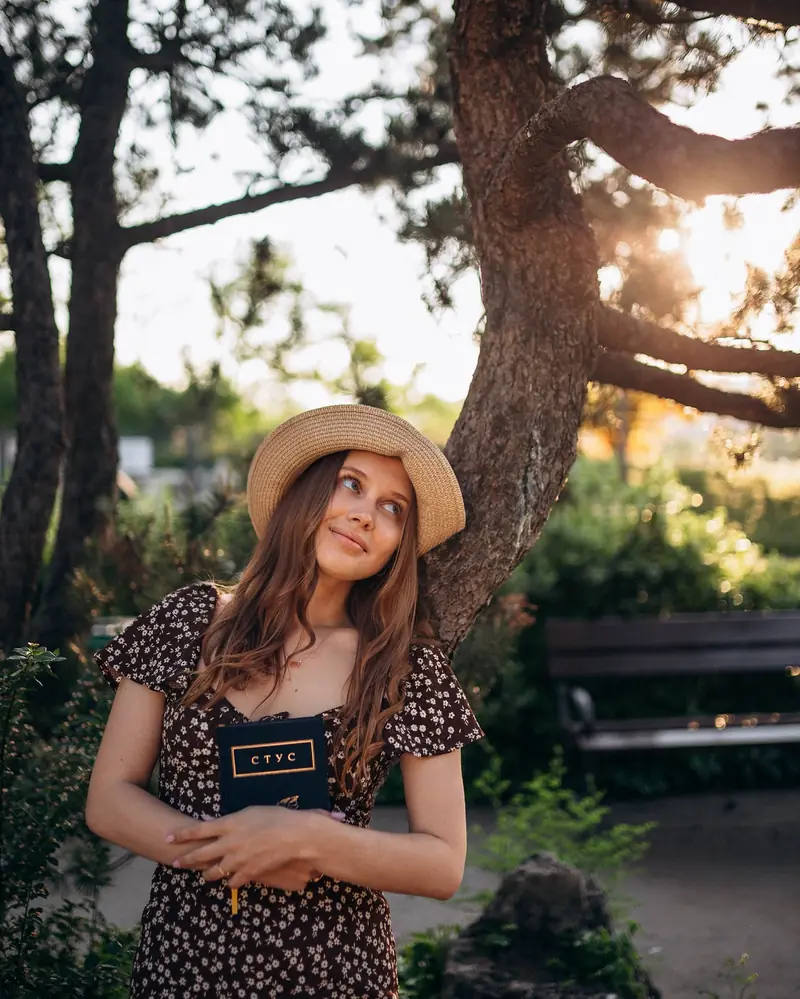 Woman in a straw hat and floral dress holding a book in a sunlit Budapest garden.
