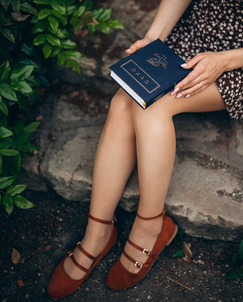 Detail of a book resting on her lap, red shoes and a floral dress on a garden step.