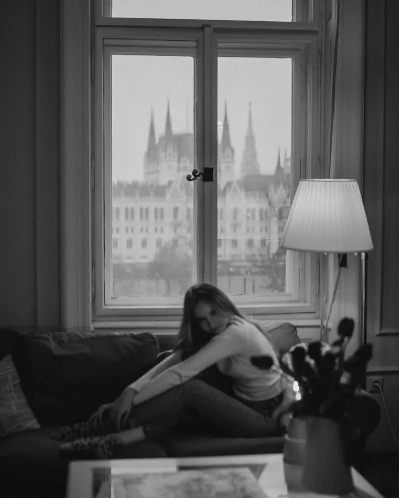 Black-and-white frame of a woman by an apartment window with the Parliament's spires outside.